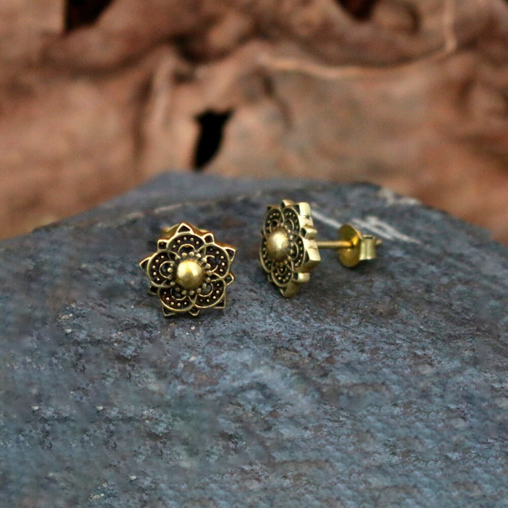 Close-up of a pair of intricate, Zenaya Brass Ear Studs by Kaya Movement. Featuring a floral design reminiscent of mandala lotus patterns, these earrings have a central bead with petal-like details extending outward. They are displayed on a textured rock surface with a blurred background.